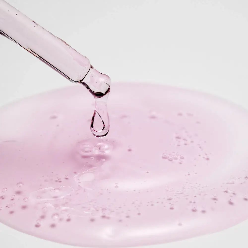 Pipette mit Tropfen rosa Flüssigkeit. Flüssigkeit auf einer Oberfläche verteilt. Keine Produktbezeichnung sichtbar.