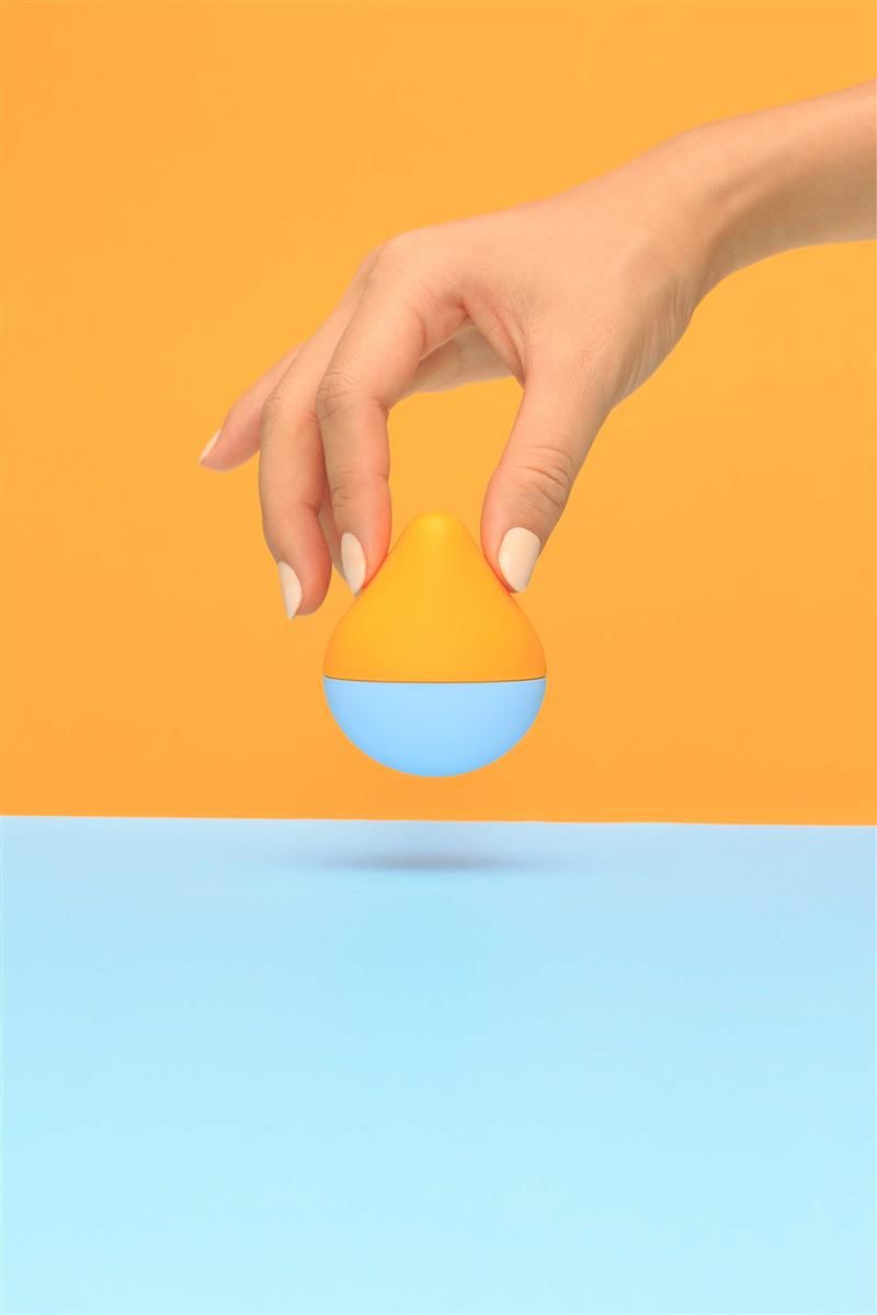 Produkt in Tropfenform, gehalten von einer Hand. Oberteil orange, Unterteil blau. Hintergrund: orange und blau. Keine weiteren Details sichtbar.
