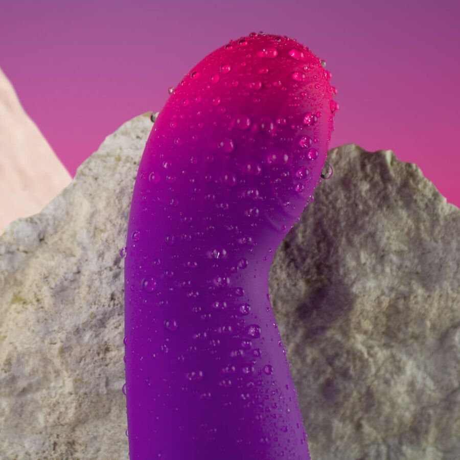 Nahaufnahme eines lila-pinkfarbenen Vibrators mit Wassertropfen. Hintergrund mit Steinen.
