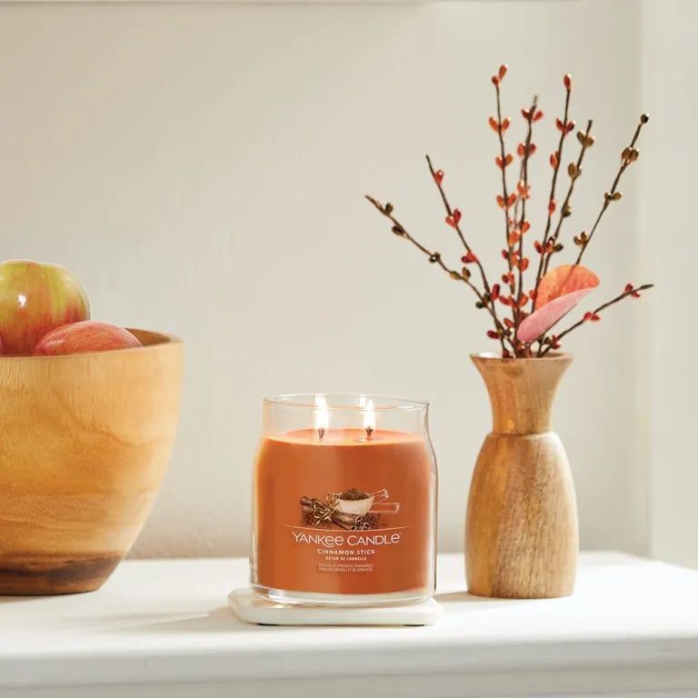 Glasbehälter mit orangefarbenem Wachs auf einem weißen Untersetzer. Daneben eine Holzschale mit Äpfeln und eine Vase mit Zweigen.
