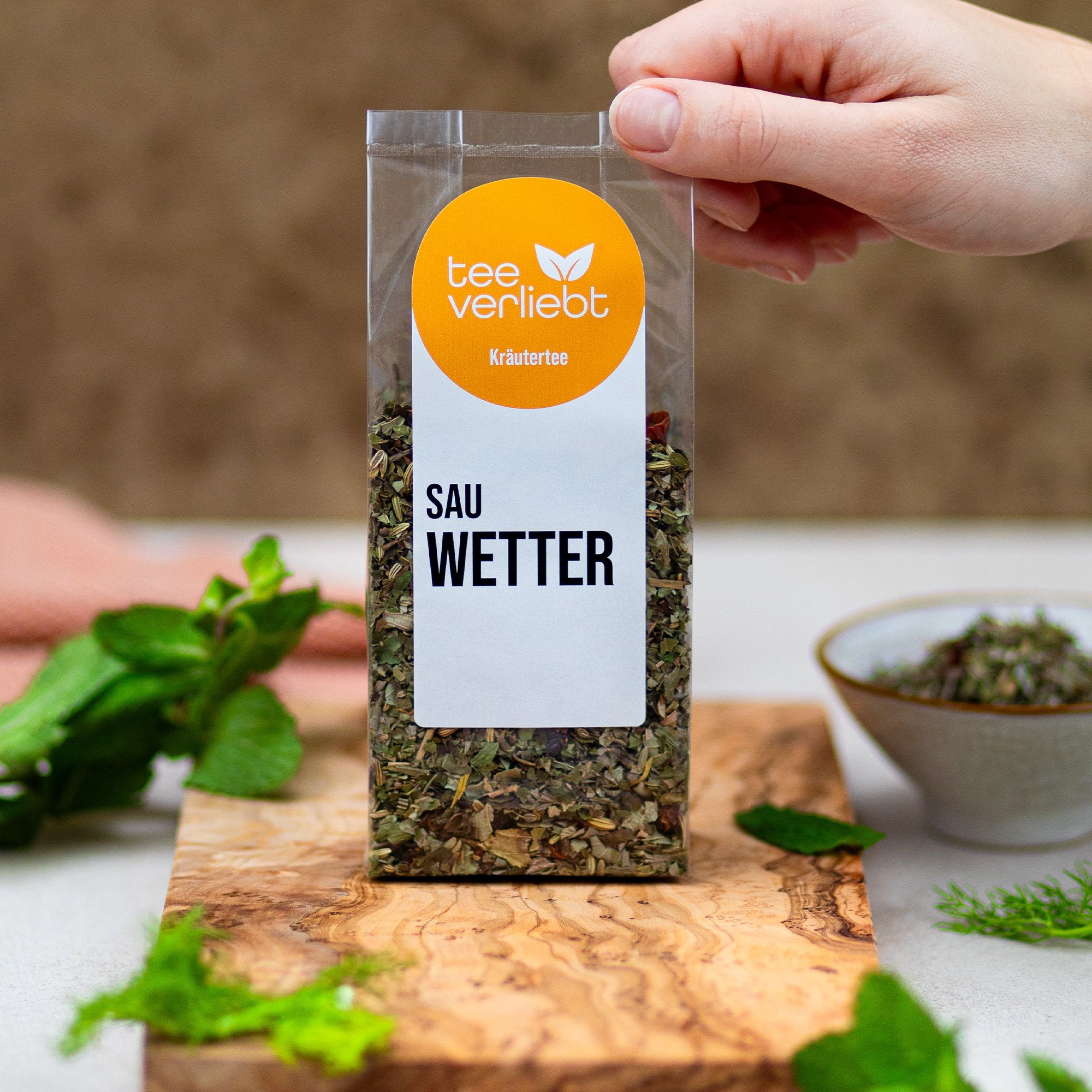 Kräutertee-Verpackung auf Holzbrett. Hand hält die Verpackung. Logo und Schriftzug "SAU WETTER" sichtbar.