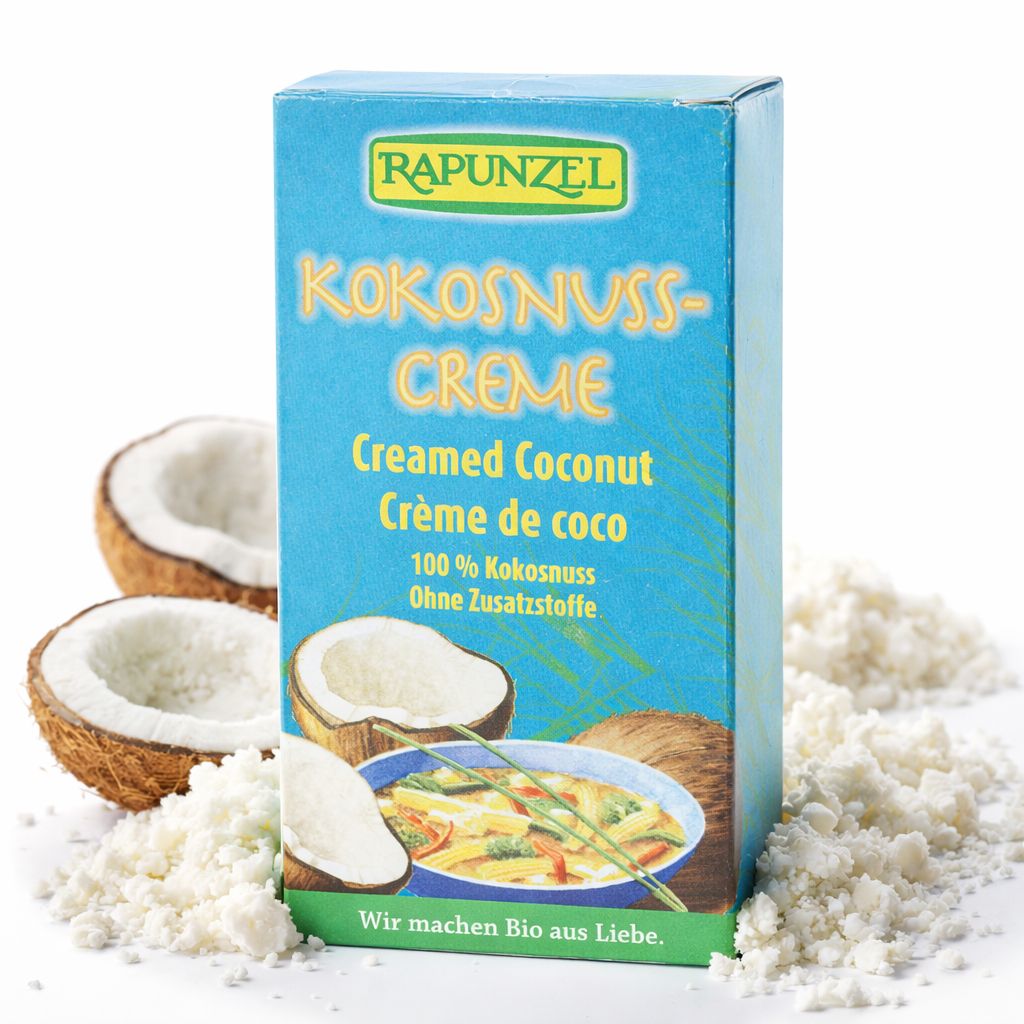 Rapunzel Kokosnuss-Creme Karton mit Kokosnuss-Hälften, Kokosraspeln und einer Schüssel Curry.