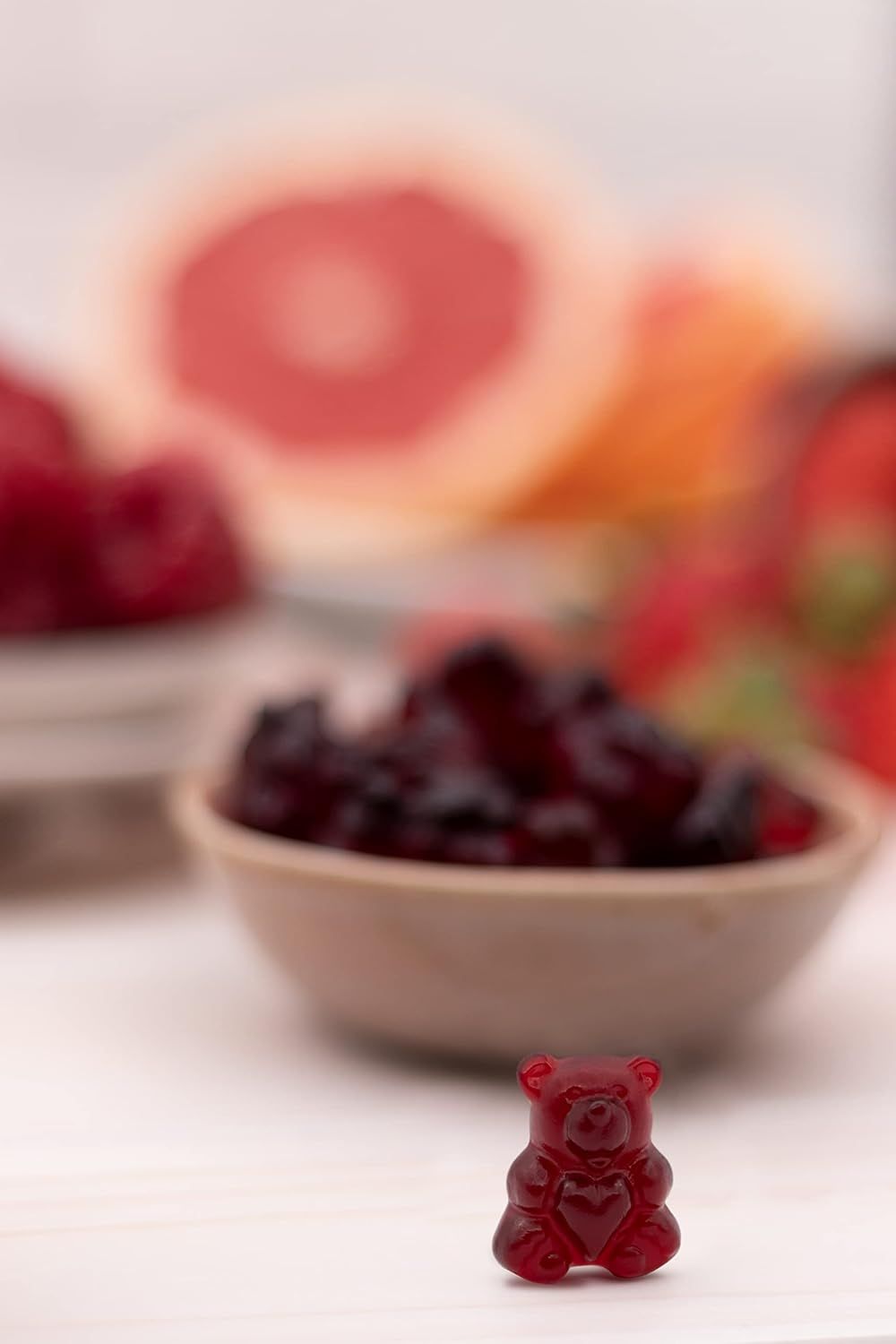 Ein roter Gummibär mit Herz. Im Hintergrund eine Schale mit weiteren Gummibären und unscharfe Früchte.