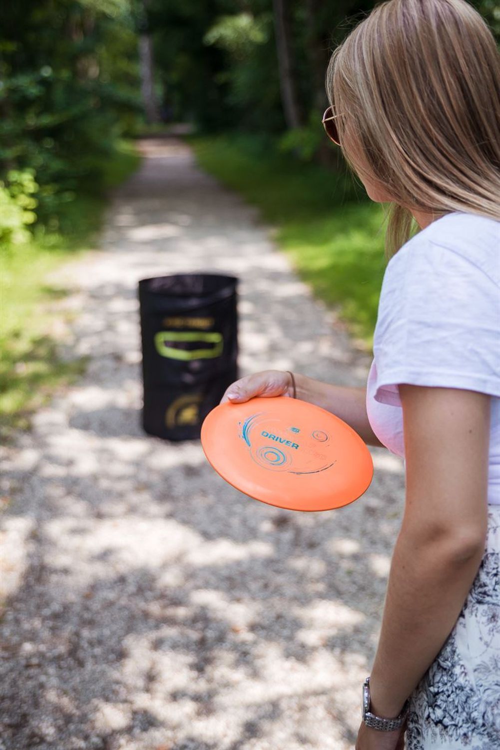 Person hält orangefarbene Disc Golf Scheibe (Driver) in der Hand. Person steht auf einem Kiesweg.