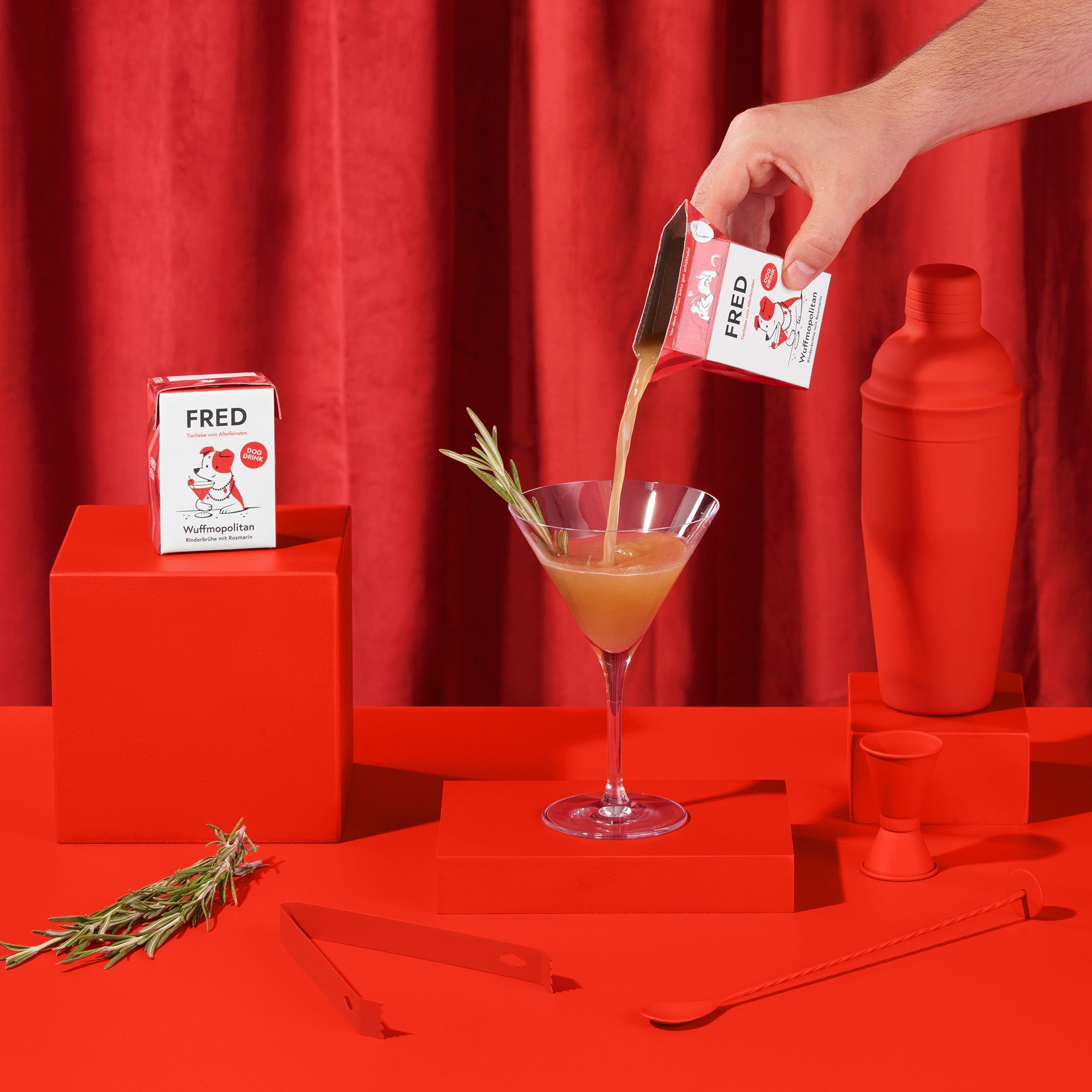 Hand gießt "FRED Dog Drink" in Cocktailglas. Roter Hintergrund. Neben: Shaker, Messbecher, Zange, Löffel, Rosmarinzweig, Würfel und eine weitere Packung.