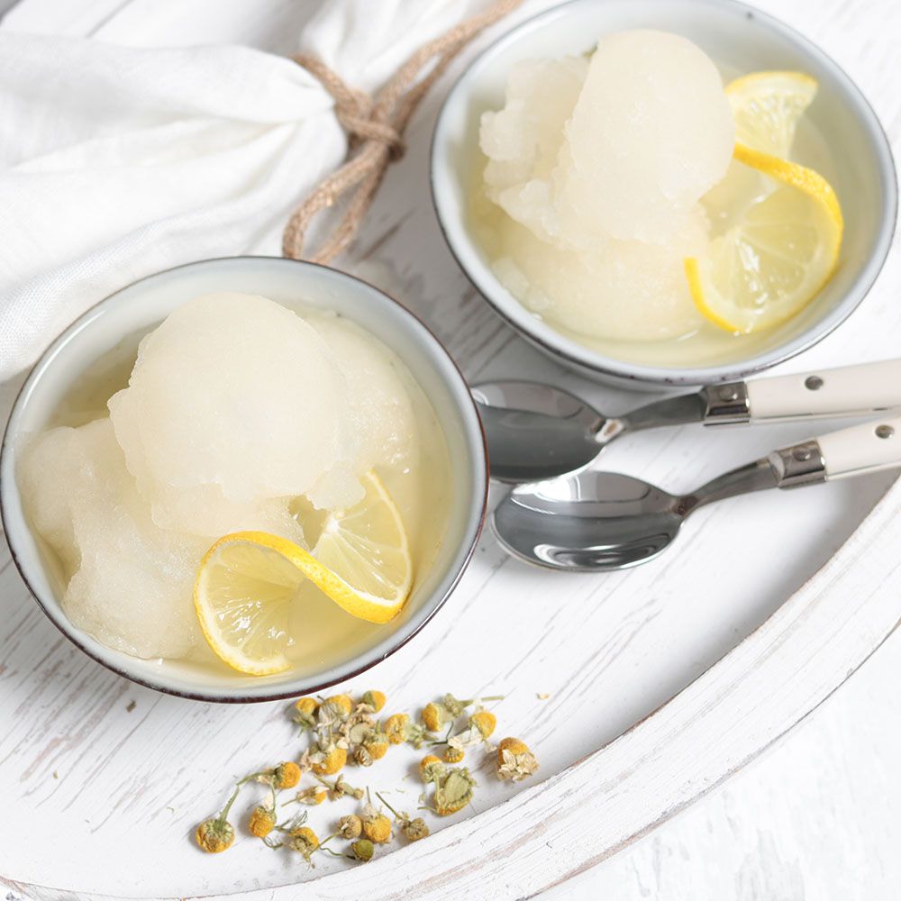 Zwei Schalen mit hellem Sorbet, Zitronenscheiben. Auf Tablett mit Löffeln und Kamillenblüten.