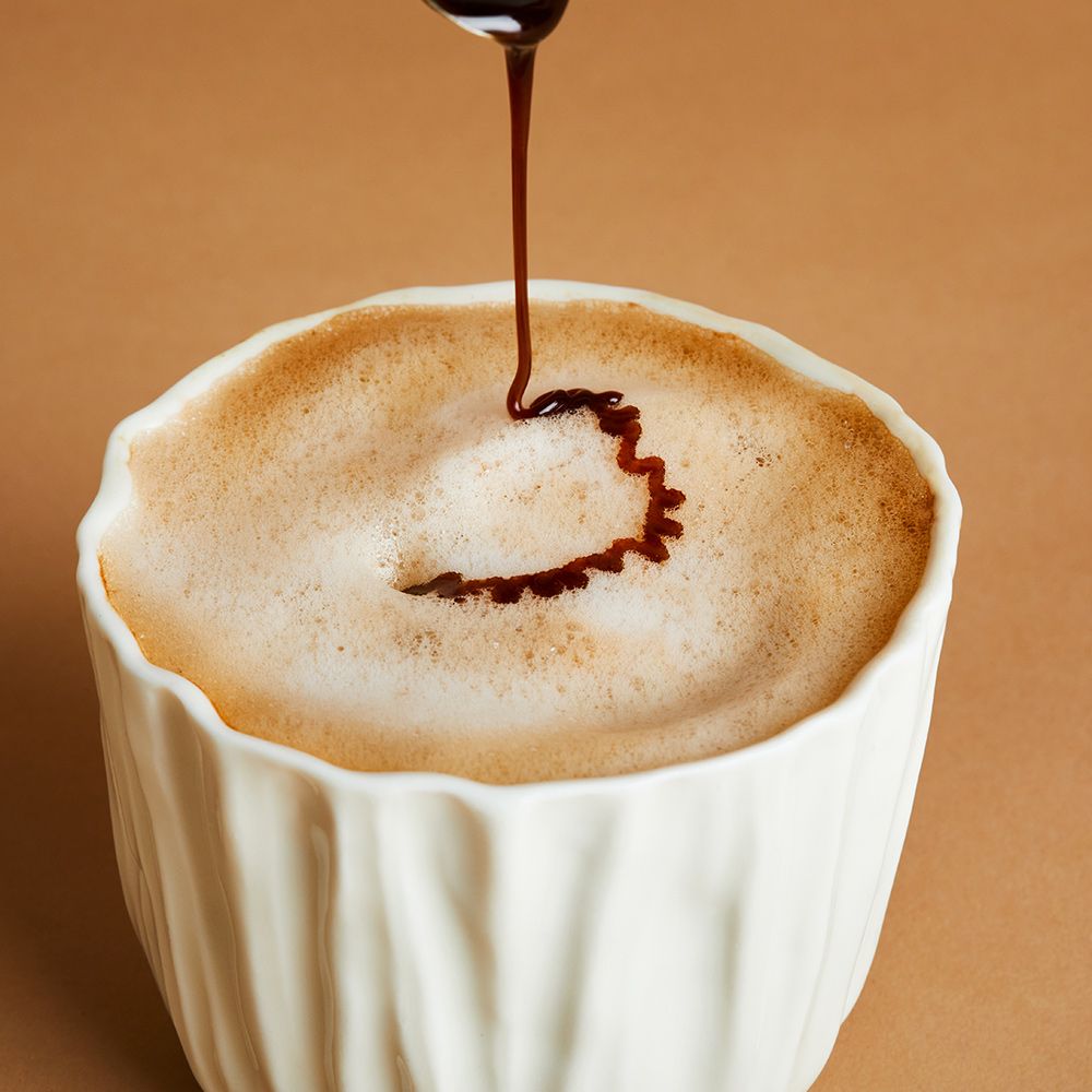 Weißes Gefäß mit Schaum, aus dem eine dunkle Flüssigkeit tropft. Der Tropfen bildet ein Muster. Hintergrund braun.