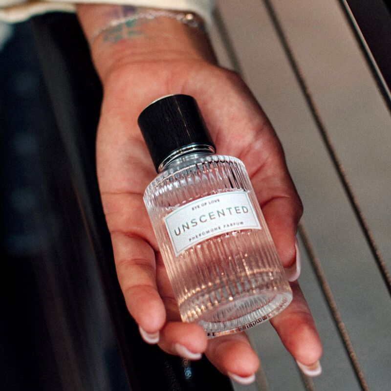Glasflakon in Hand gehalten. Aufschrift: Unscented, Eye of Love, Pheromon Parfum. Hand mit Tattoo.