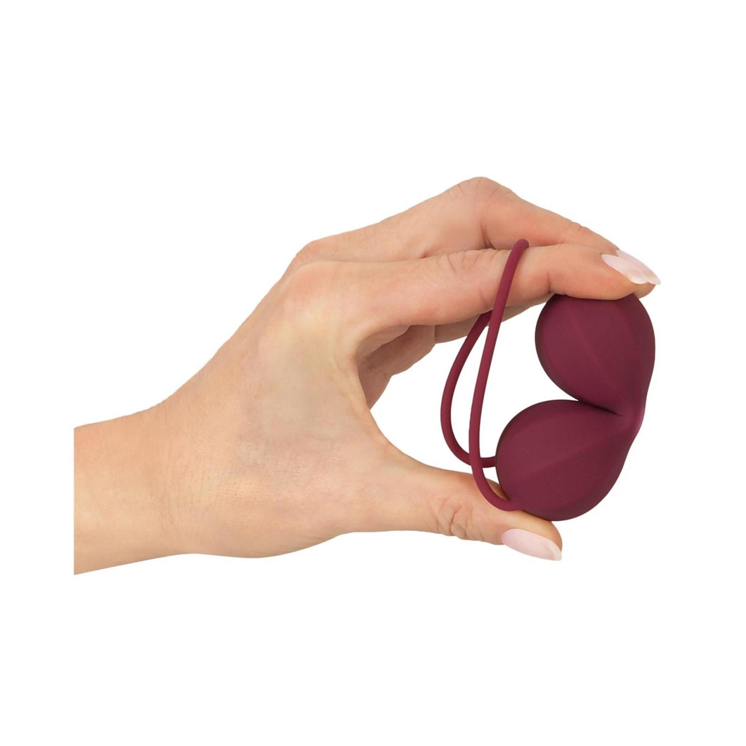 Hand hält burgunderfarbene Liebeskugel. Zwei Kugeln, gerillte Oberfläche, Schlaufe. Weißer Hintergrund.