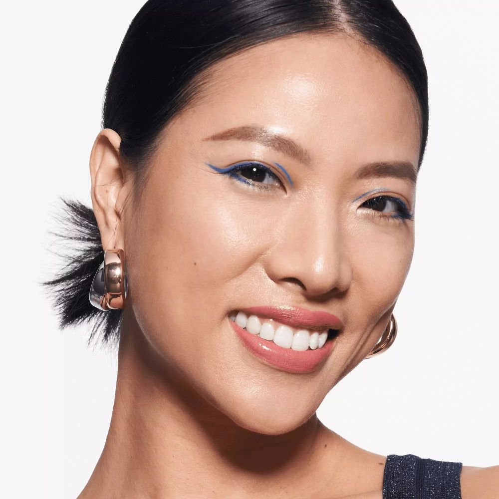 Frau mit blauem Lidstrich. Trägt Ohrringe. Lächelt.