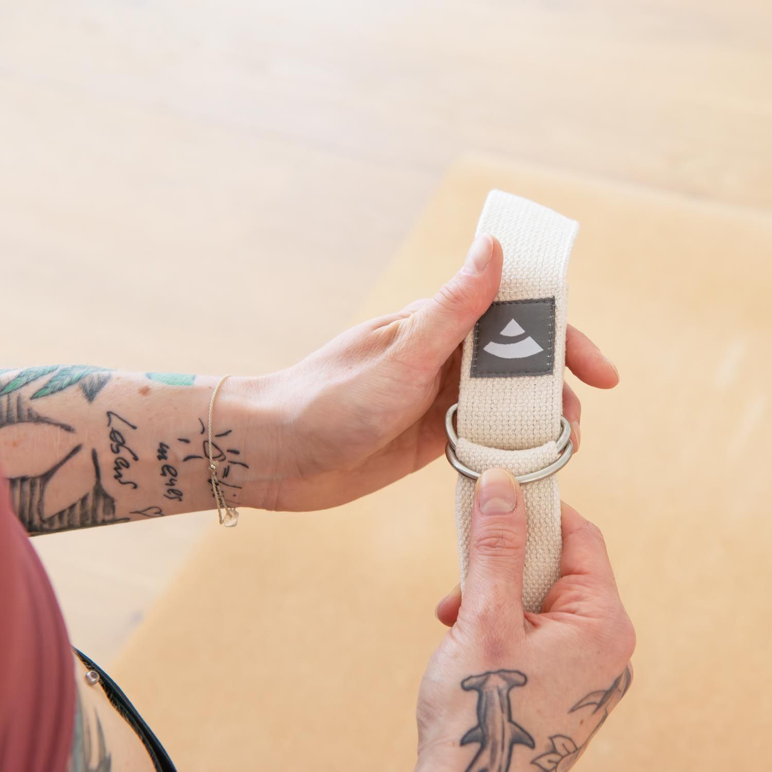 Person hält einen beigen Yogagurt. Metallring und Logo sind sichtbar.