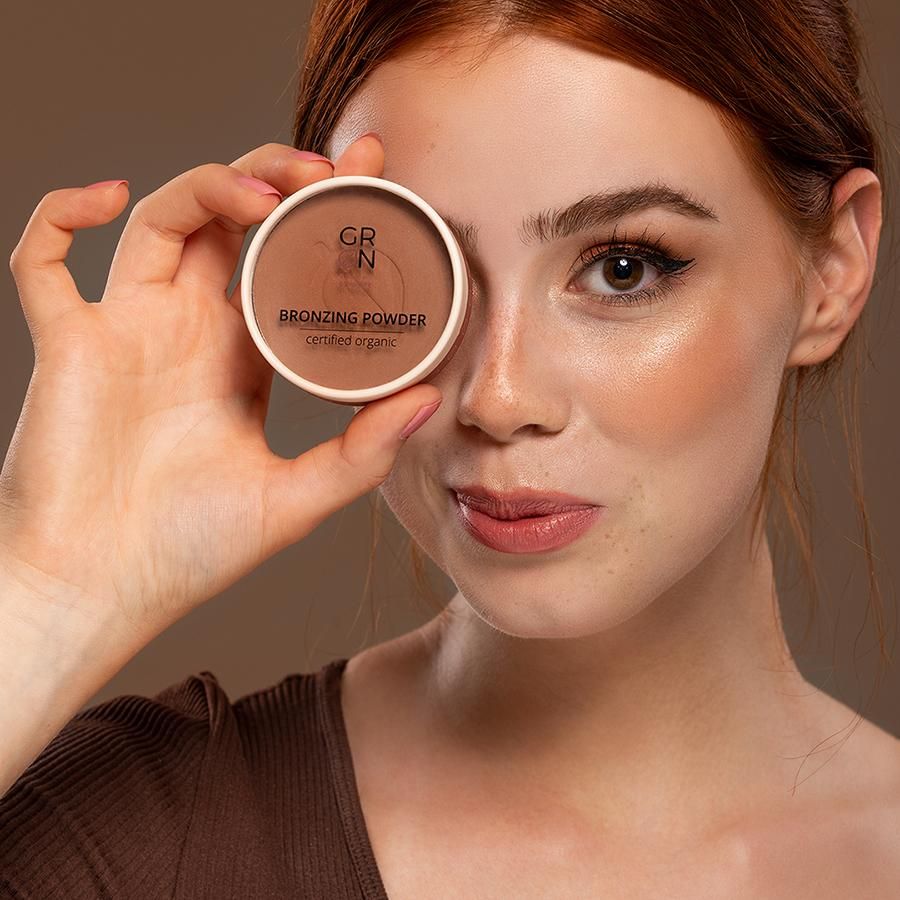Frau hält ein rundes Döschen Bronzing Powder vor das Auge. Aufschrift: GRN Bronzing Powder certified organic.