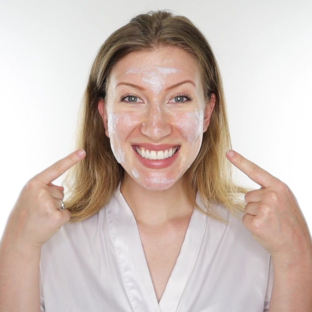 Frau mit weißer Creme im Gesicht. Sie zeigt mit den Fingern auf ihr Gesicht. Sie trägt einen weißen Bademantel.