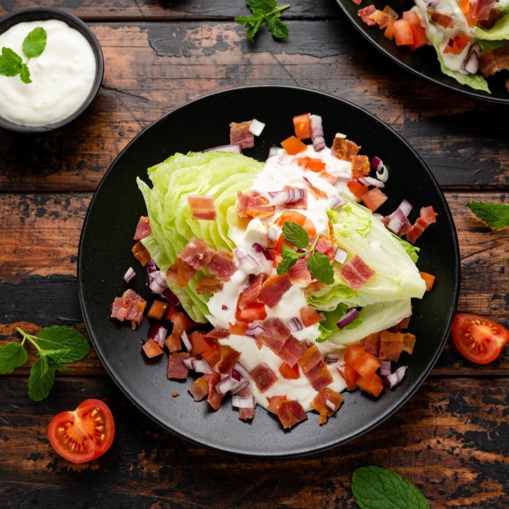 Salat auf schwarzem Teller. Eisbergsalat, Tomaten, Speck und Sauce. Minze als Dekoration. Auf Holzuntergrund.