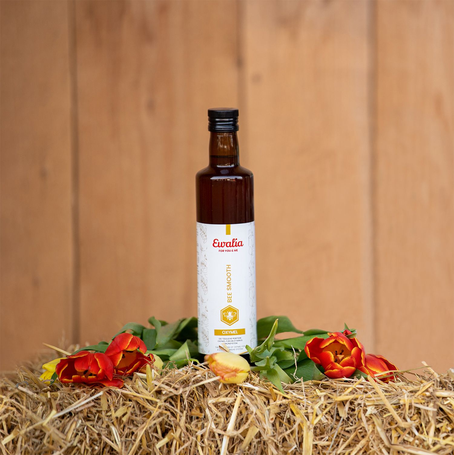 Braune Glasflasche mit Etikett. Auf Heuballen mit roten Tulpen und Blättern. Aufschrift: Ewalia, BEE SMOOTH, OXYMEL.