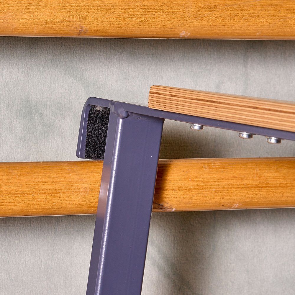 Detailaufnahme des Sprossenwandsitzes. Holzplatte, Metallrahmen, schwarze Polsterung. Rahmen an Sprossenwand befestigt.