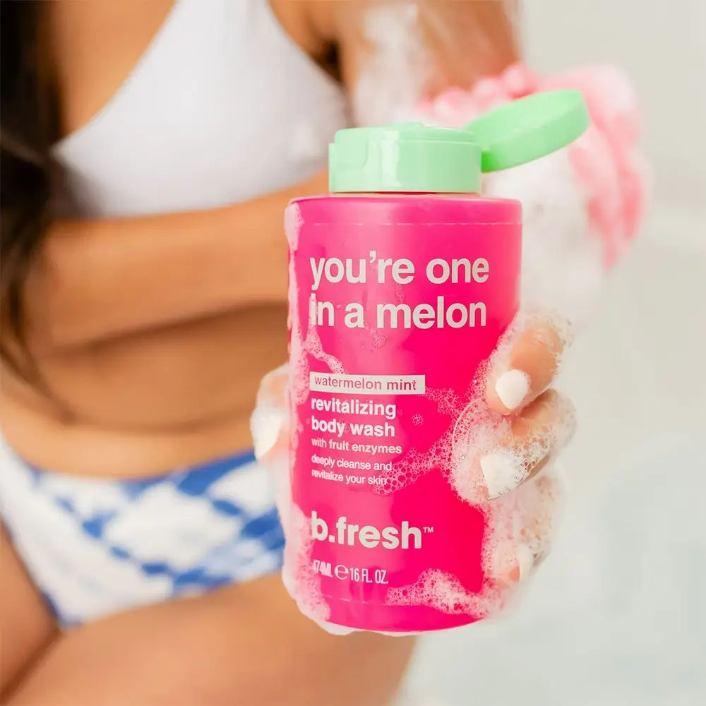 Rosa Duschgel-Flasche in Hand, umgeben von Schaum. Grüner Deckel. Text: "you're one in a melon", "b.fresh". Person im Hintergrund.