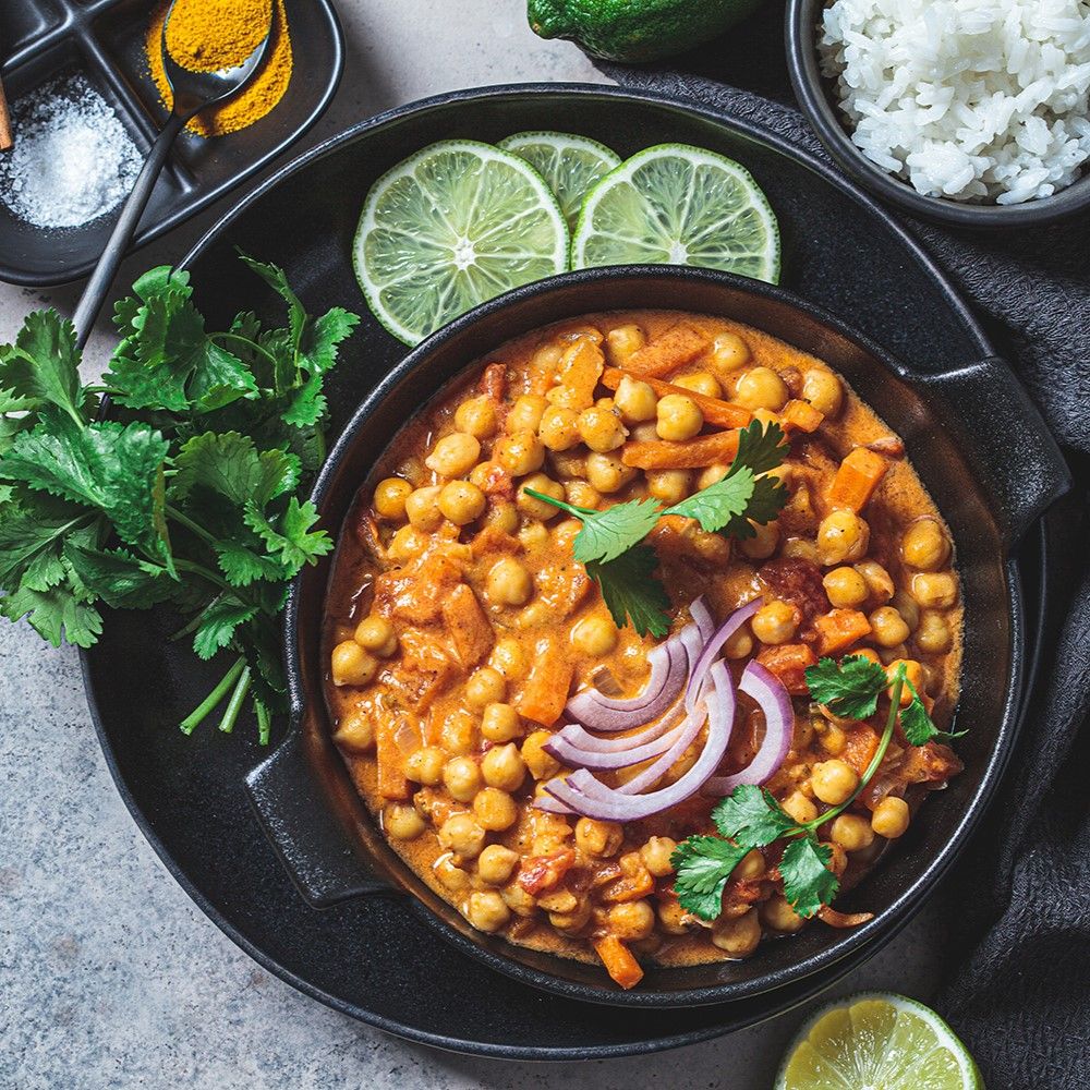 Kichererbsen-Curry in schwarzer Schüssel, garniert mit Zwiebeln und Koriander. Mit Reis, Limettenscheiben und Gewürzen.