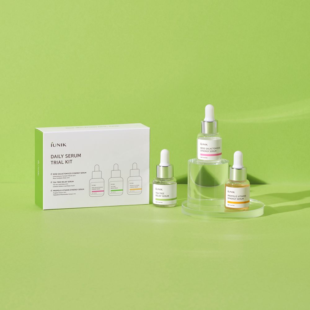 Weiße Verpackung und drei Serumflaschen auf einer transparenten Plattform vor grünem Hintergrund. Auf der Verpackung steht "IUNIK DAILY SERUM TRIAL KIT".