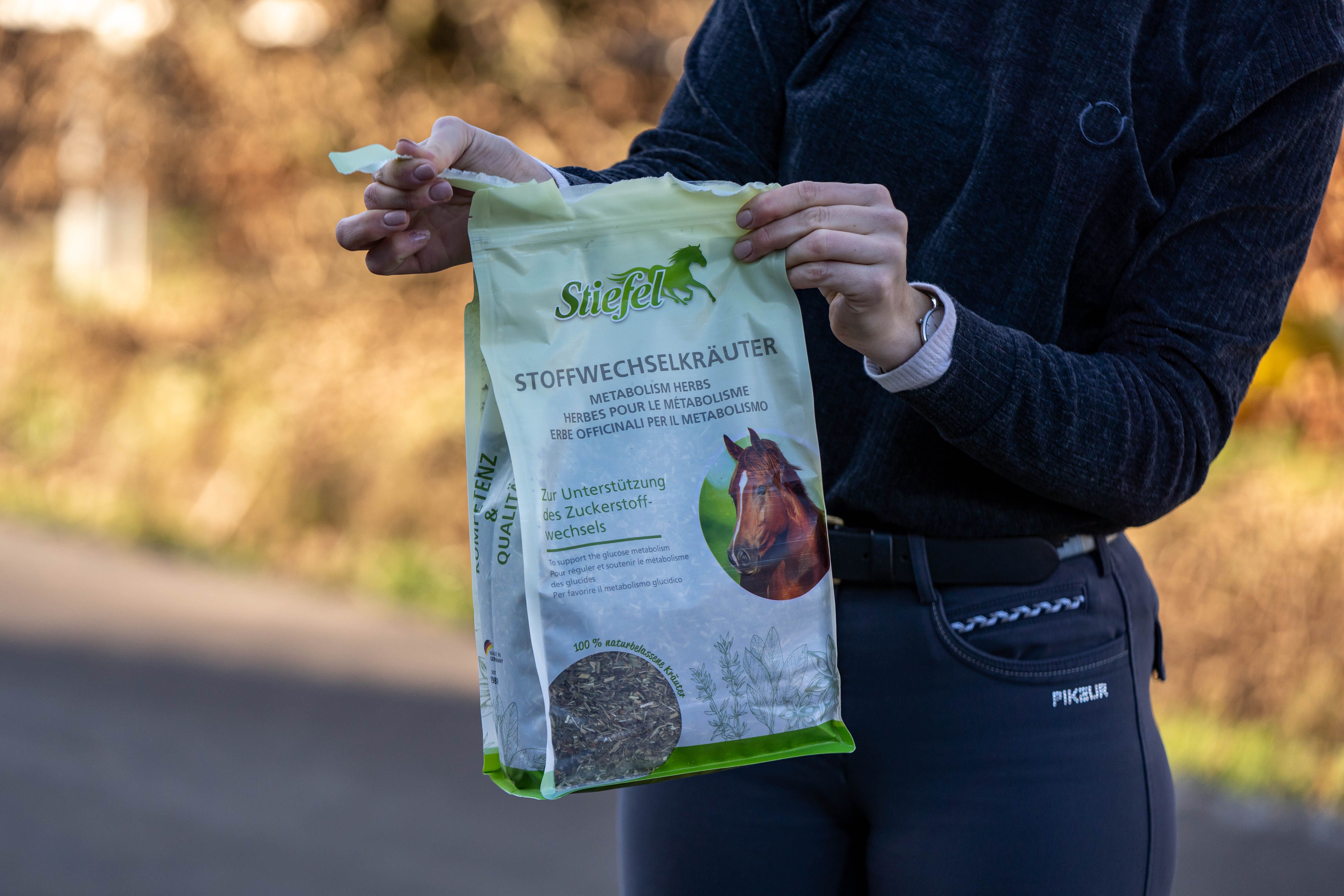 Person hält eine grüne Verpackung mit Produktnamen. Produktname und Logo deutlich sichtbar. Hintergrund: verschwommene Landschaft.