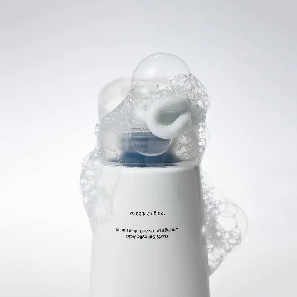 Weiße Tube mit Schaum. Text: 0.5% Salicylsäure. 120 g / 4.23 oz. Schaum um den Ausgießer.