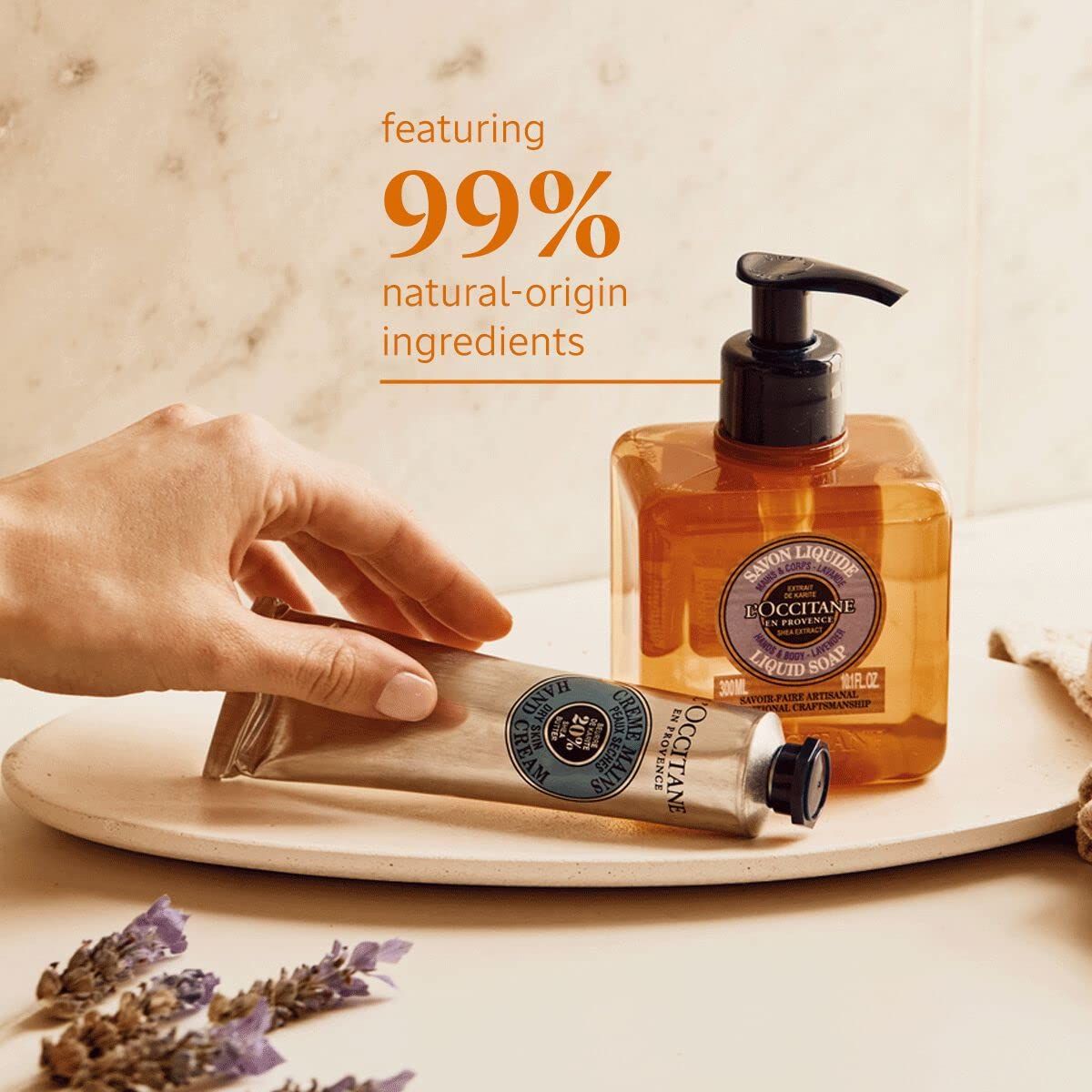 Flüssigseife und Handcreme auf Tablett. Hand berührt Flasche. Text: L'Occitane, 99% natürliche Inhaltsstoffe.
