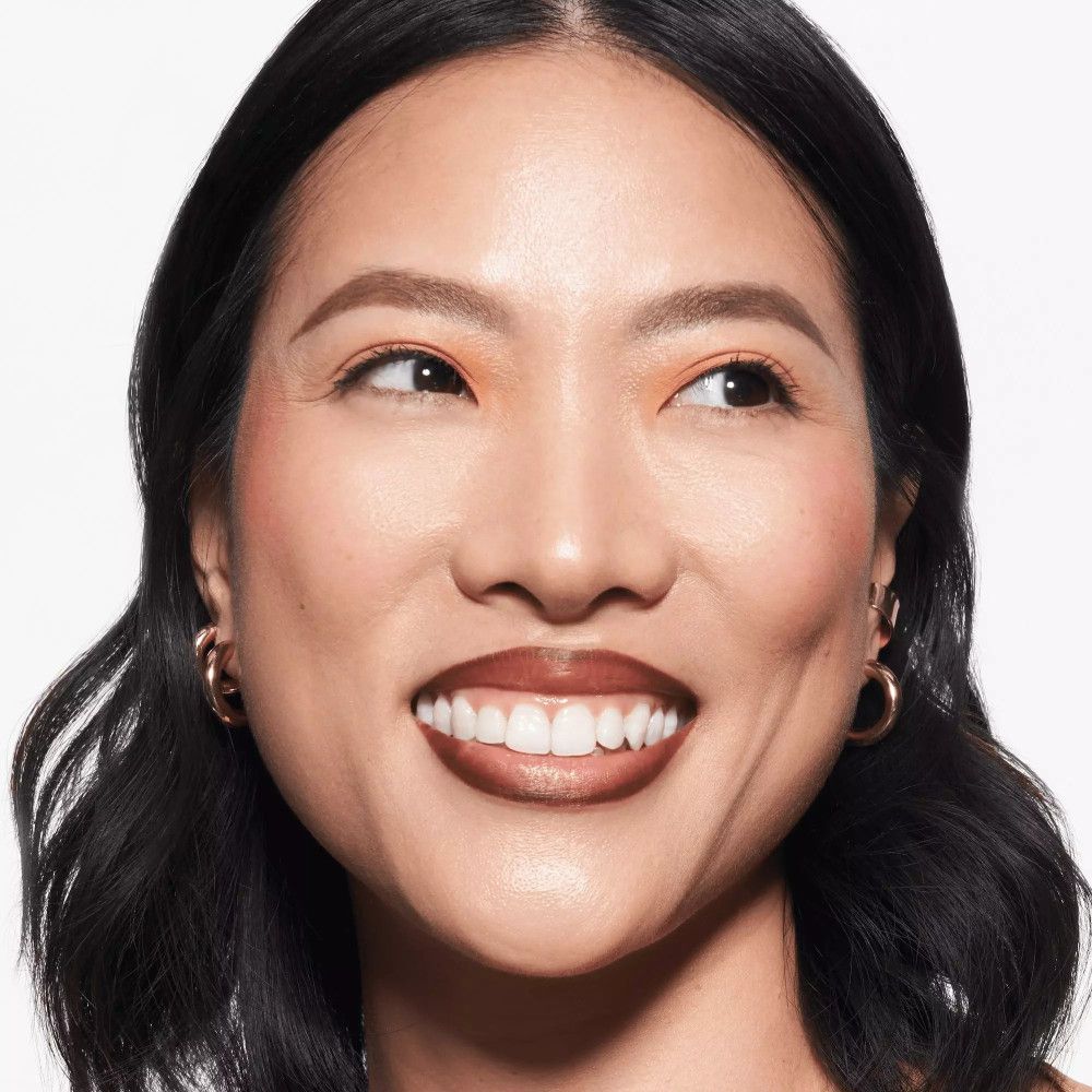 Porträt einer lächelnden Frau mit braunem Lippenstift. Geformte Augenbrauen, goldene Ohrringe.