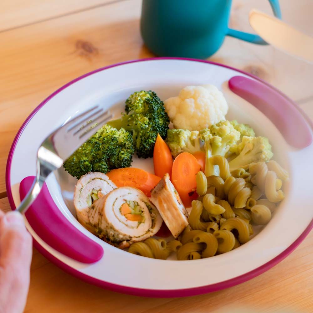 Teller mit rosa Griffen, gefüllt mit Gemüse und Fleisch. Gabel in der Hand. Auf einem Holztisch.