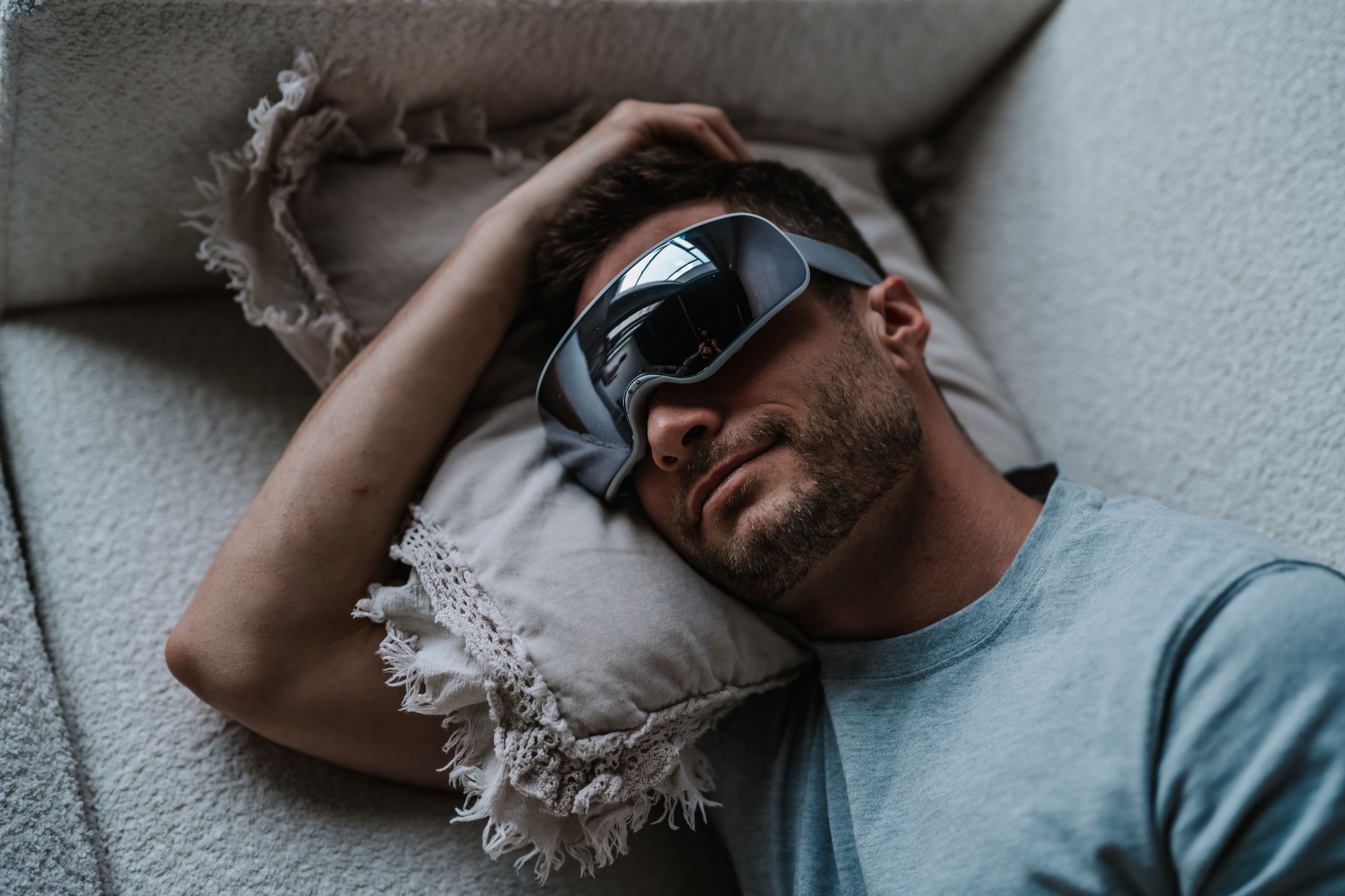 Mann liegt auf dem Rücken, Augenmassagegerät auf den Augen. Graues Gerät, graues Band. Auf Kissen und Decke.