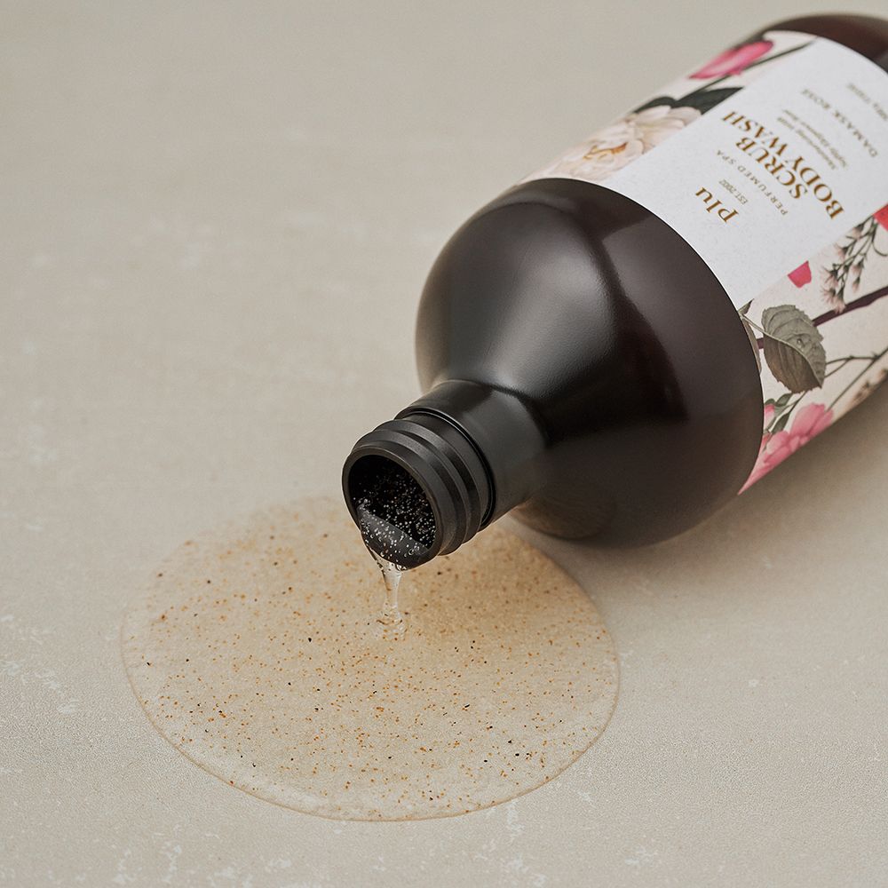 Braune Flasche mit schwarzem Pumpverschluss, aus der Flüssigkeit austritt. Etikett mit floralem Design und Schriftzug: Scrub Body Wash, Damask Rose. Marke: plu.