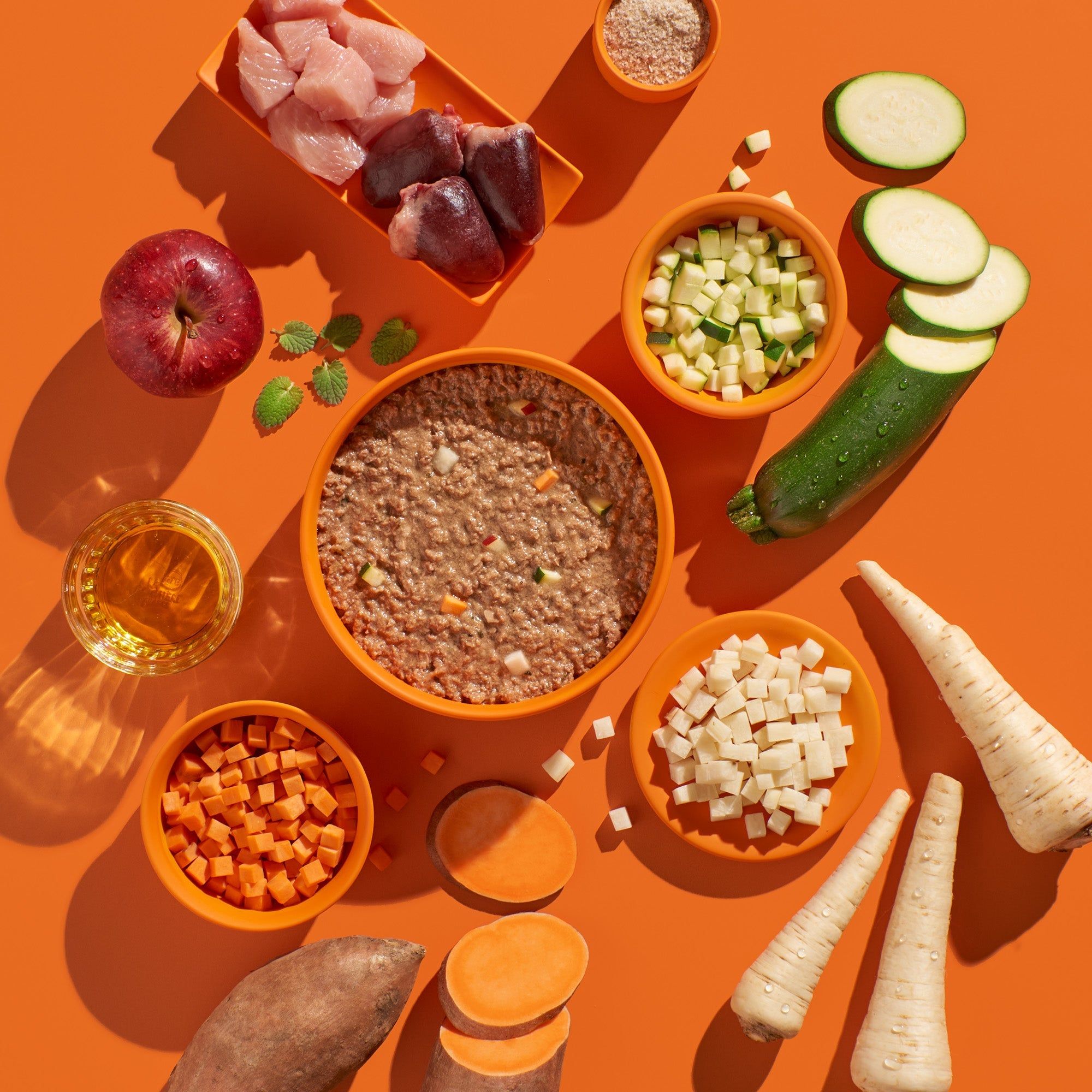 Verschiedene Zutaten in Schalen auf orangefarbenem Hintergrund: Fleisch, Gemüse, Obst. Zucchini, Pastinaken, Süßkartoffeln, Apfel.