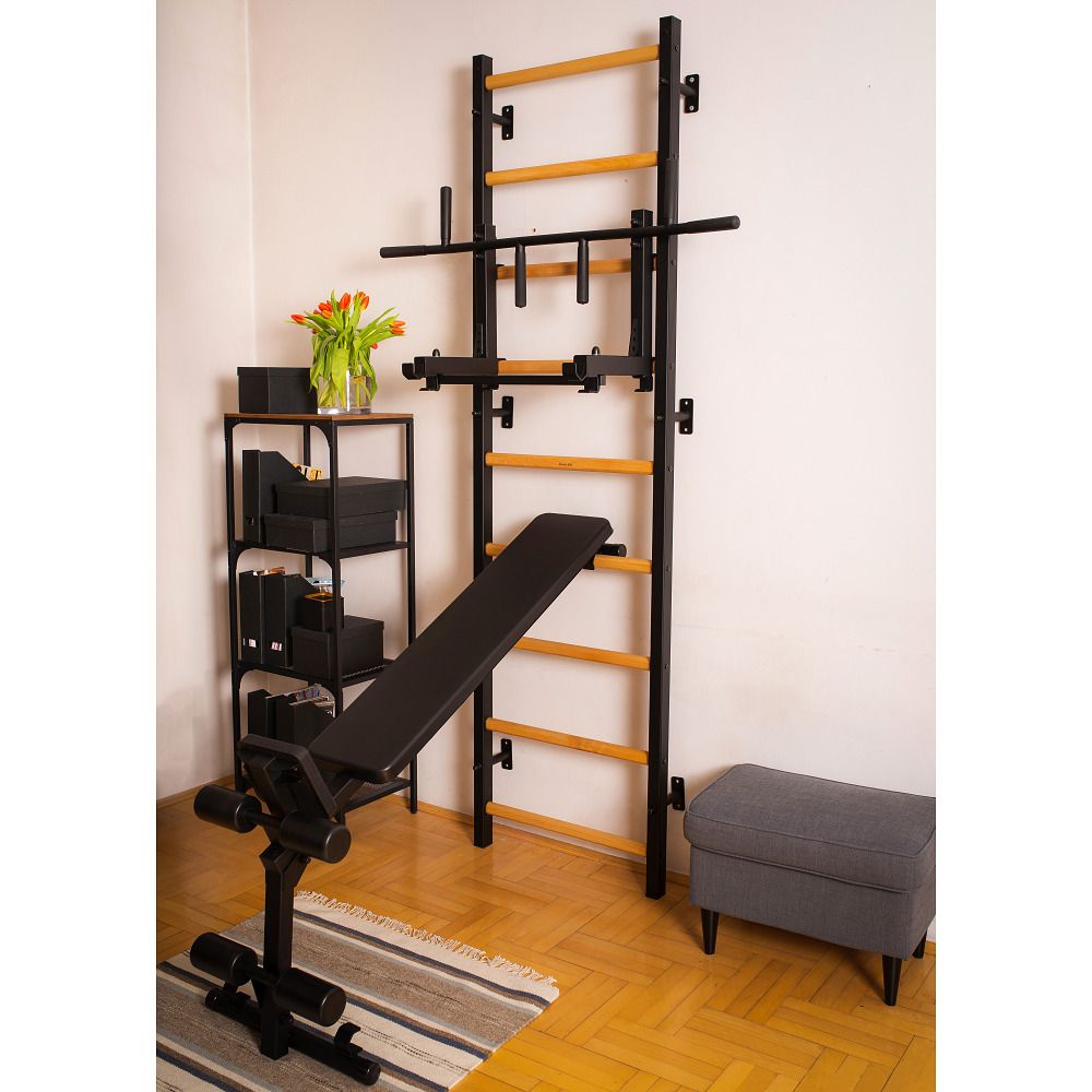 Fitness-Sprossenwand mit Zubehör. Eine schwarze Bank steht vor der Wand. Blumen und ein Regal sind zu sehen.