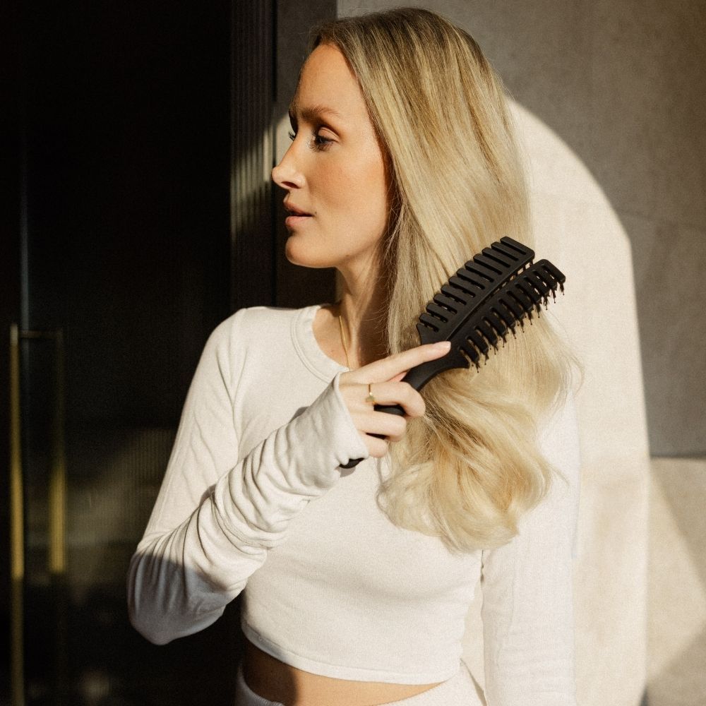 Frau kämmt sich mit schwarzer Haarbürste die Haare. Blondes Haar.