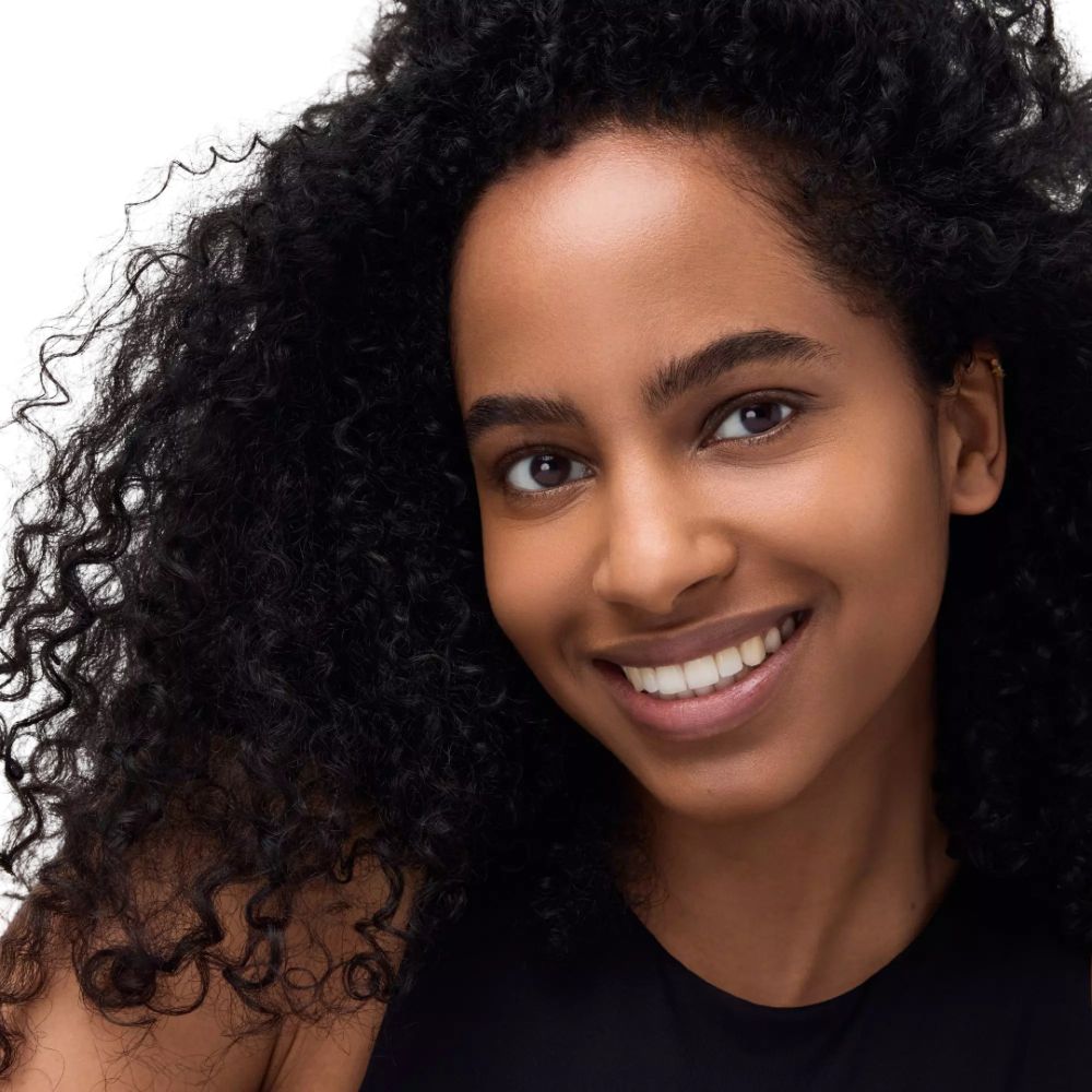 Porträt einer Frau mit lockigem Haar. Sie lächelt. Schwarzes Top. Weißer Hintergrund.