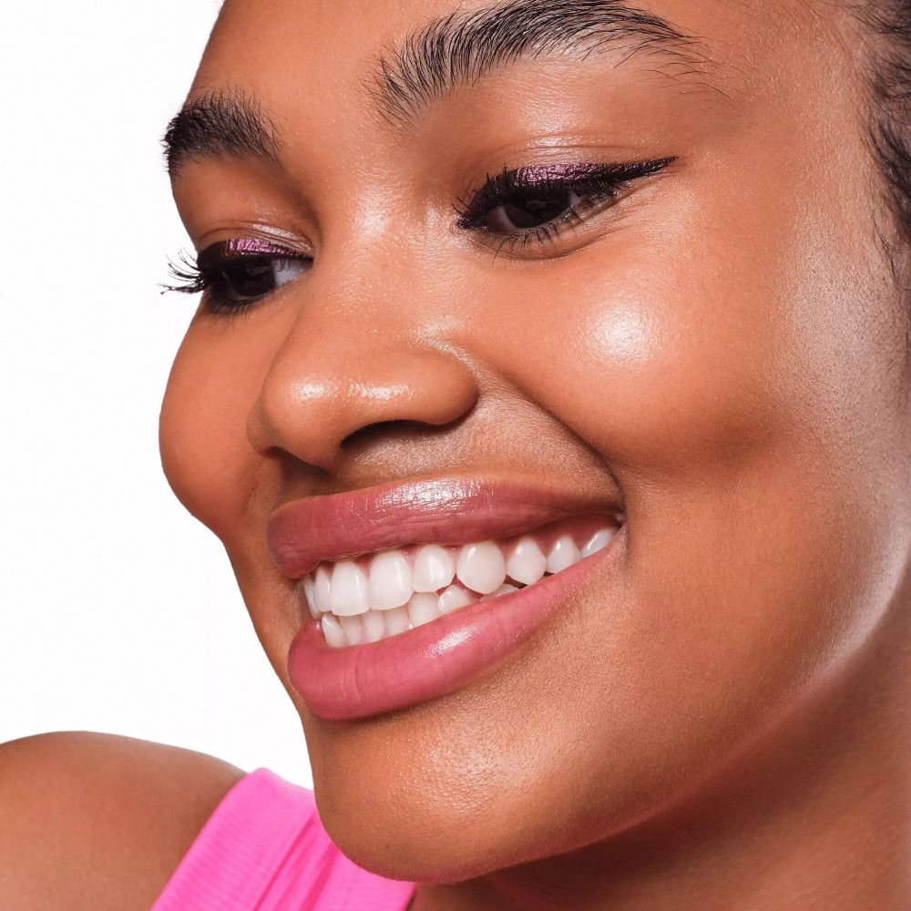 Porträt einer Frau mit lila Metallic-Eyeliner. Die Frau hat dunkle Haare und trägt ein rosa Top. Sie lächelt.