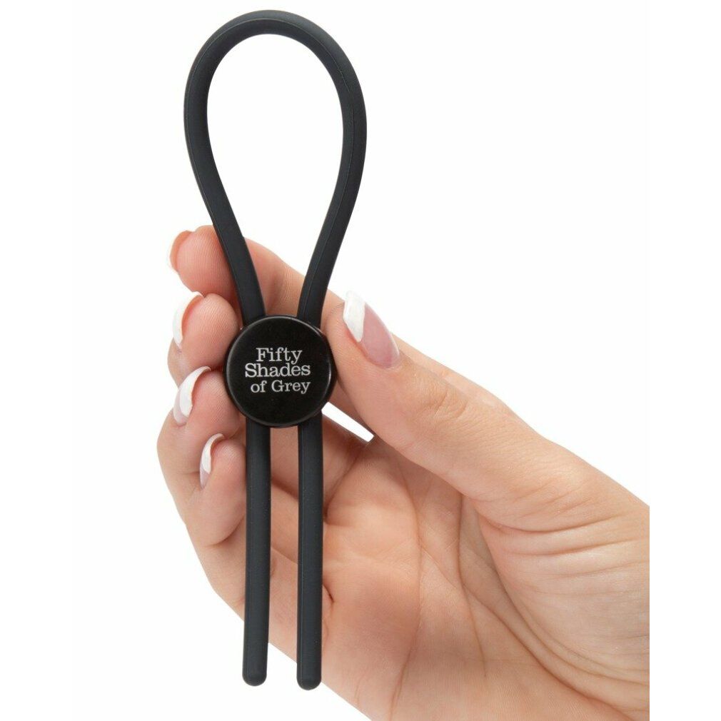 Penisschlaufe in Hand gehalten. Dunkelgrau, rundes Mittelteil mit Schriftzug. Zwei Stränge und ein Ring.