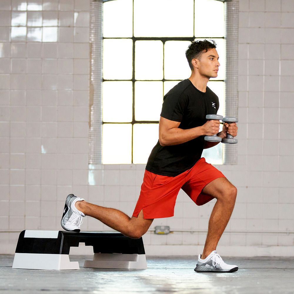 Mann trainiert mit Hanteln auf einem Reebok Step. Er trägt Sportkleidung und Schuhe. Der Step ist schwarz-weiß.