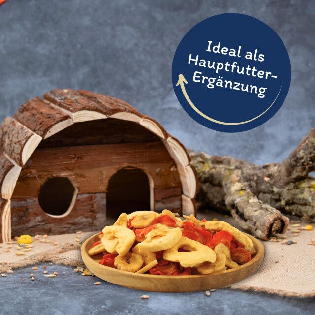 Getrocknete Obst- und Gemüsestücke in einer Holzschale. Text: 'Ideal als Hauptfutter-Ergänzung'. Hintergrund: Holzhäuschen und Ast.