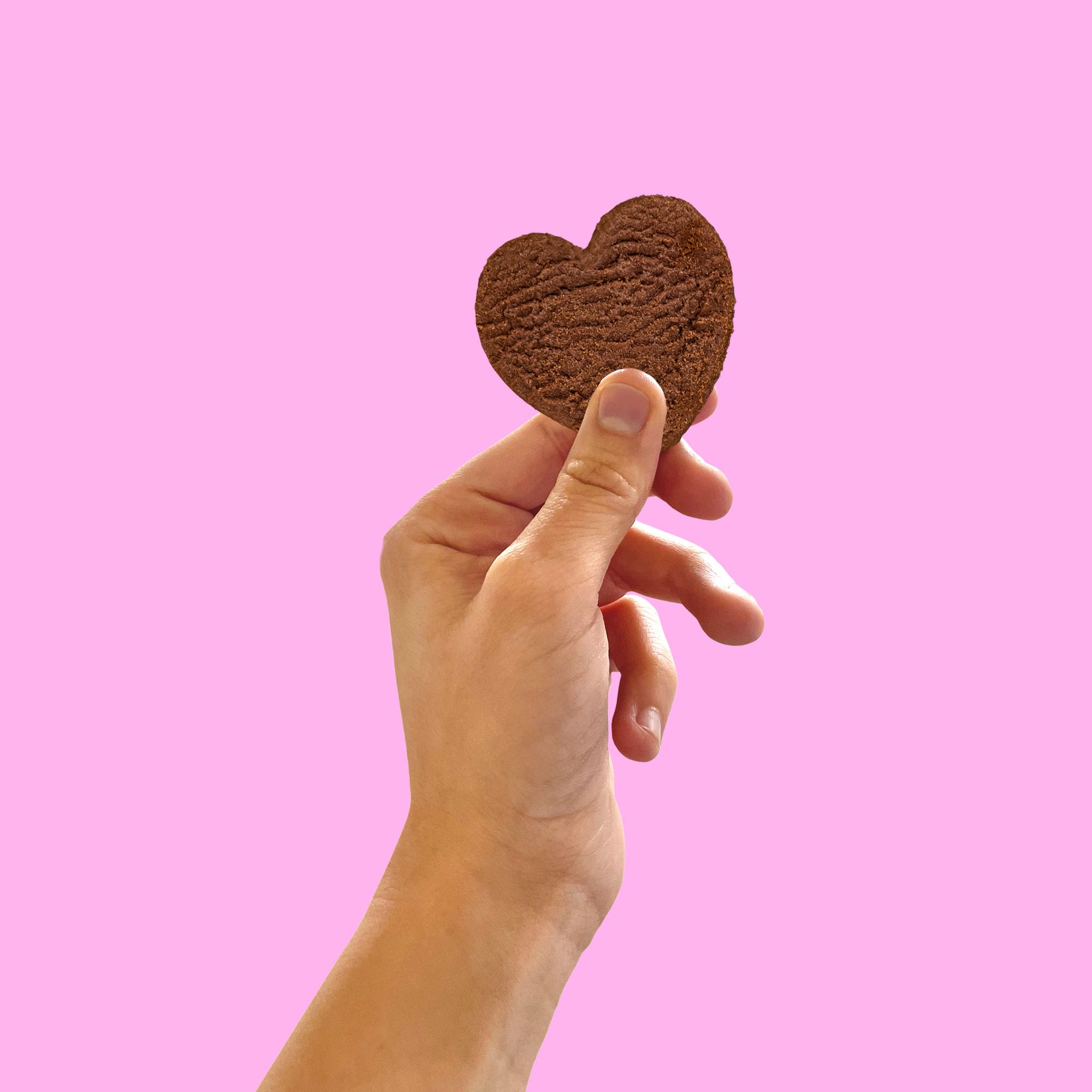 Hand hält einen IoBoscoVivo glutenfreien Kakaokeks in Herzform vor rosa Hintergrund.