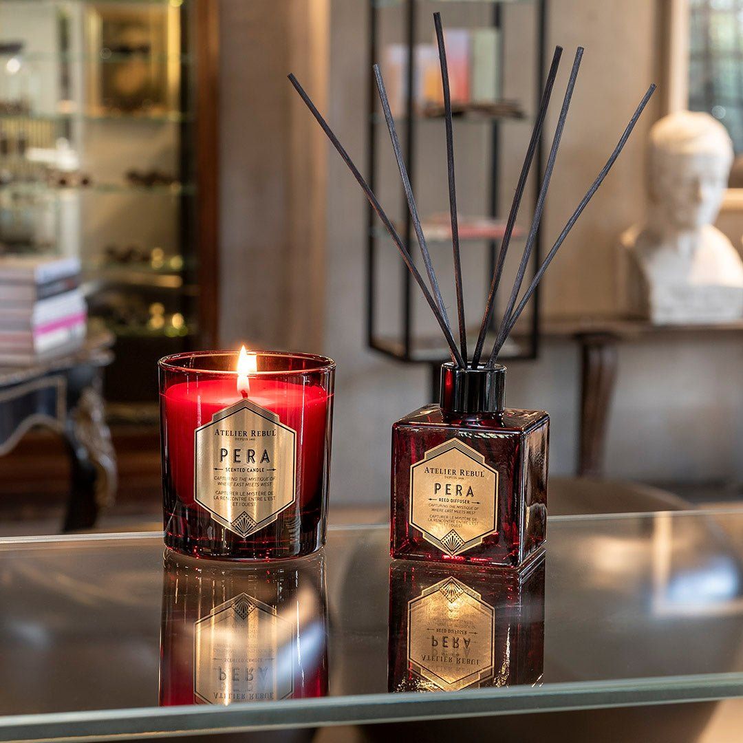 Roter Glasbehälter mit Kerze und Duftstäbchen. Beide mit Etikett 'Pera'. Schwarze Stäbe stecken in quadratischer Flasche.