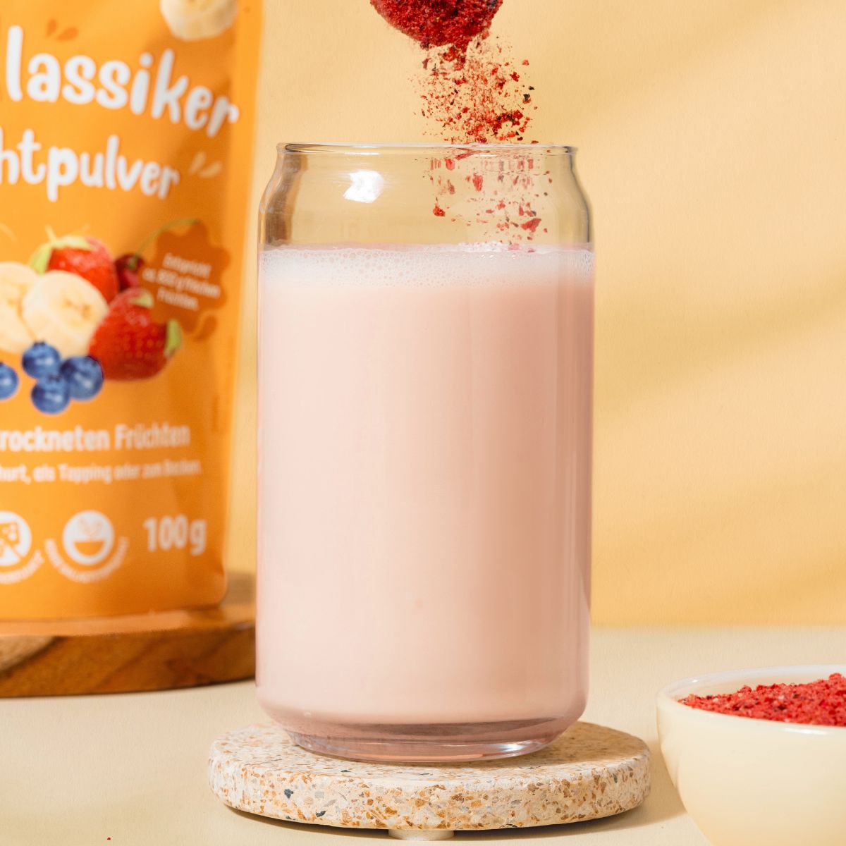 Glas mit rosa Flüssigkeit, Fruchtpulver wird hineingegossen. Daneben eine Tüte mit Fruchtpulver und eine Schale mit Fruchtpulver.