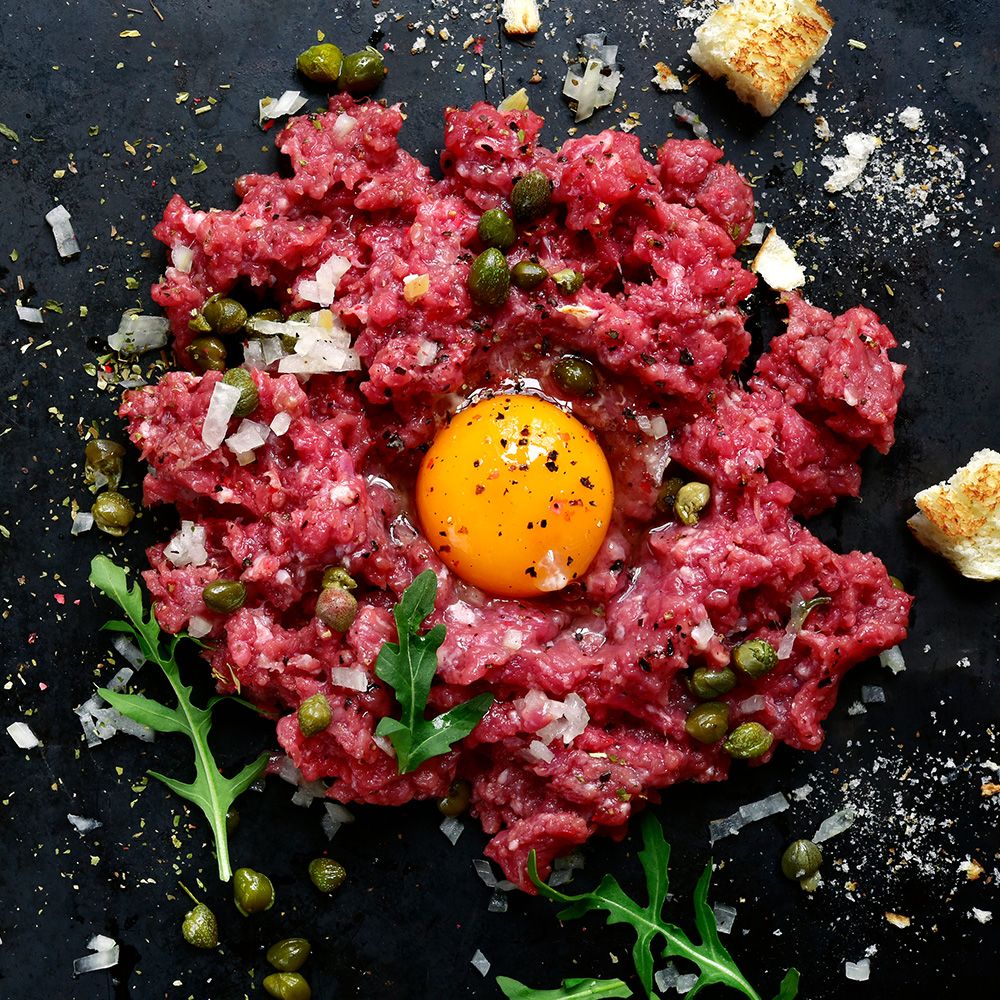 Rindertartar mit Eigelb, Kapern, Zwiebeln, Rucola und Brot. Auf schwarzem Untergrund angerichtet.