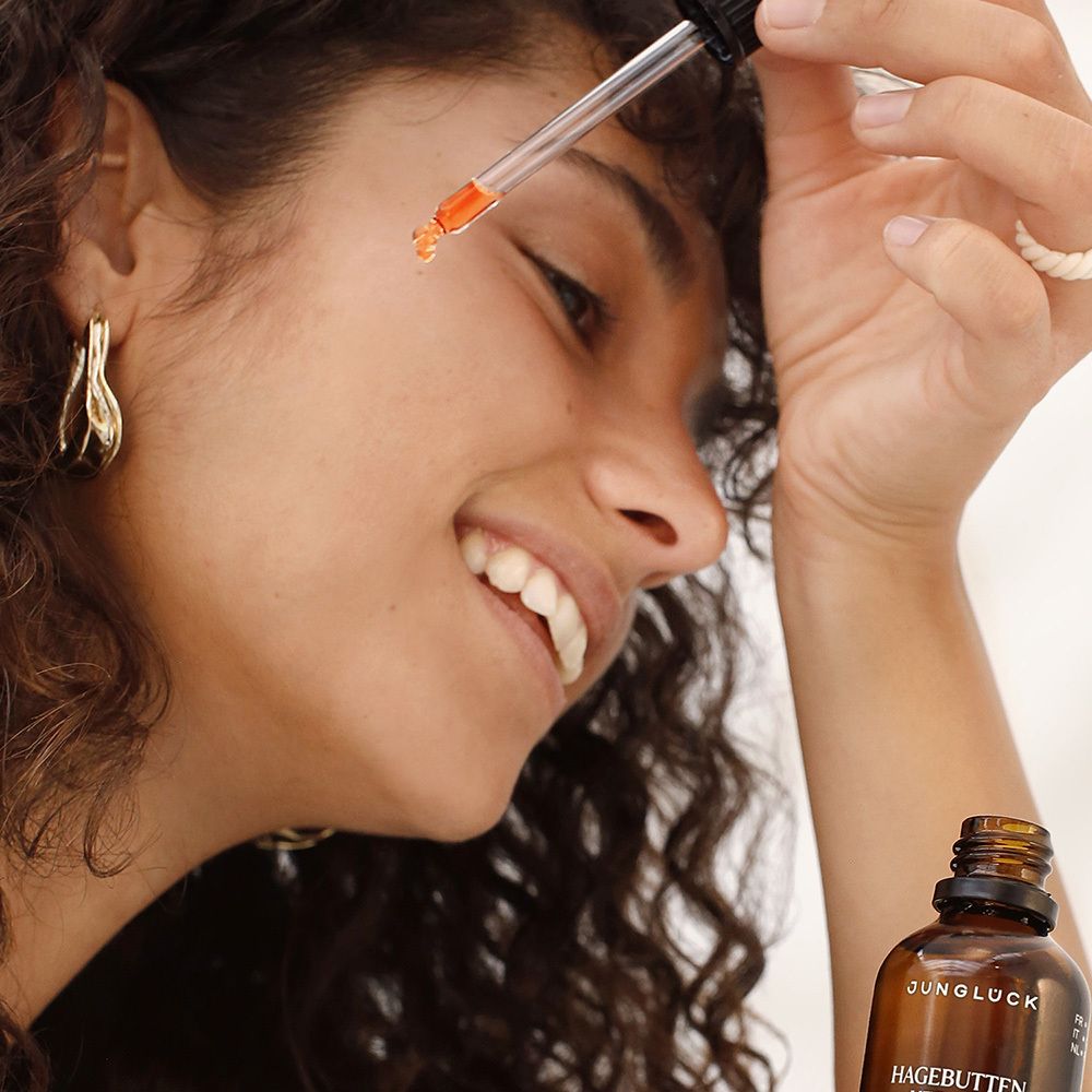 Frau mit Locken hält Pipette mit orangefarbenem Öl. Im Hintergrund: Flasche mit Aufschrift JUNGLÜCK Hagebuttenkernöl.