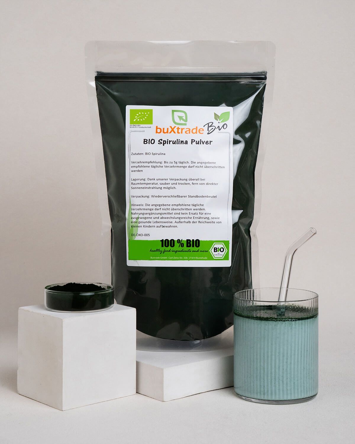Buxtrade Bio Spirulinapulver, ein Glas mit grünem Getränk und Strohhalm, kleiner Behälter mit Pulver.