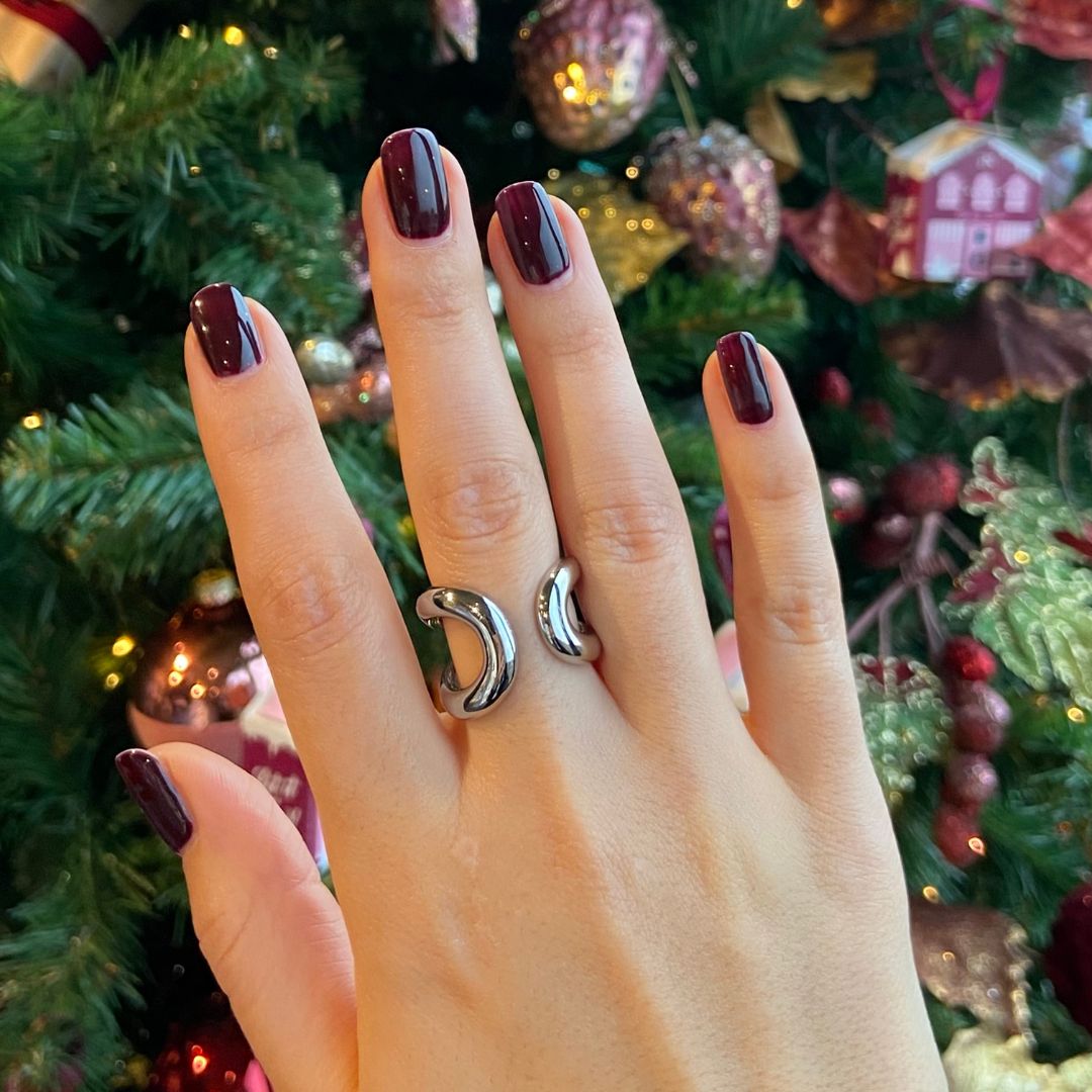 Hand mit lackierten, dunkelroten Nägeln. Die Nägel sind kurz und glänzend. Ein silberner Ring ist sichtbar. Weihnachtsbaum im Hintergrund.