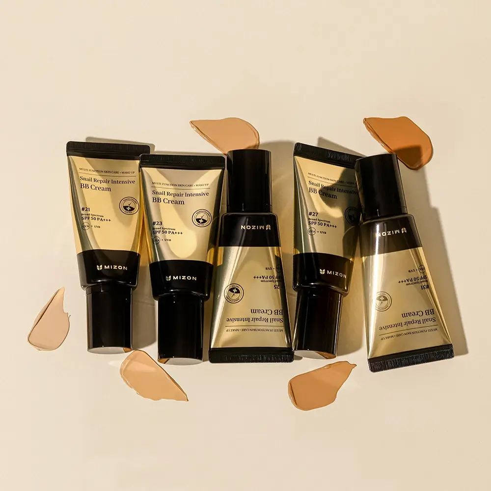 Fünf Tuben BB Cream, Marke MIZON, mit Farbmustern. Goldfarbene Tuben mit schwarzem Deckel. Auf hellem Untergrund.