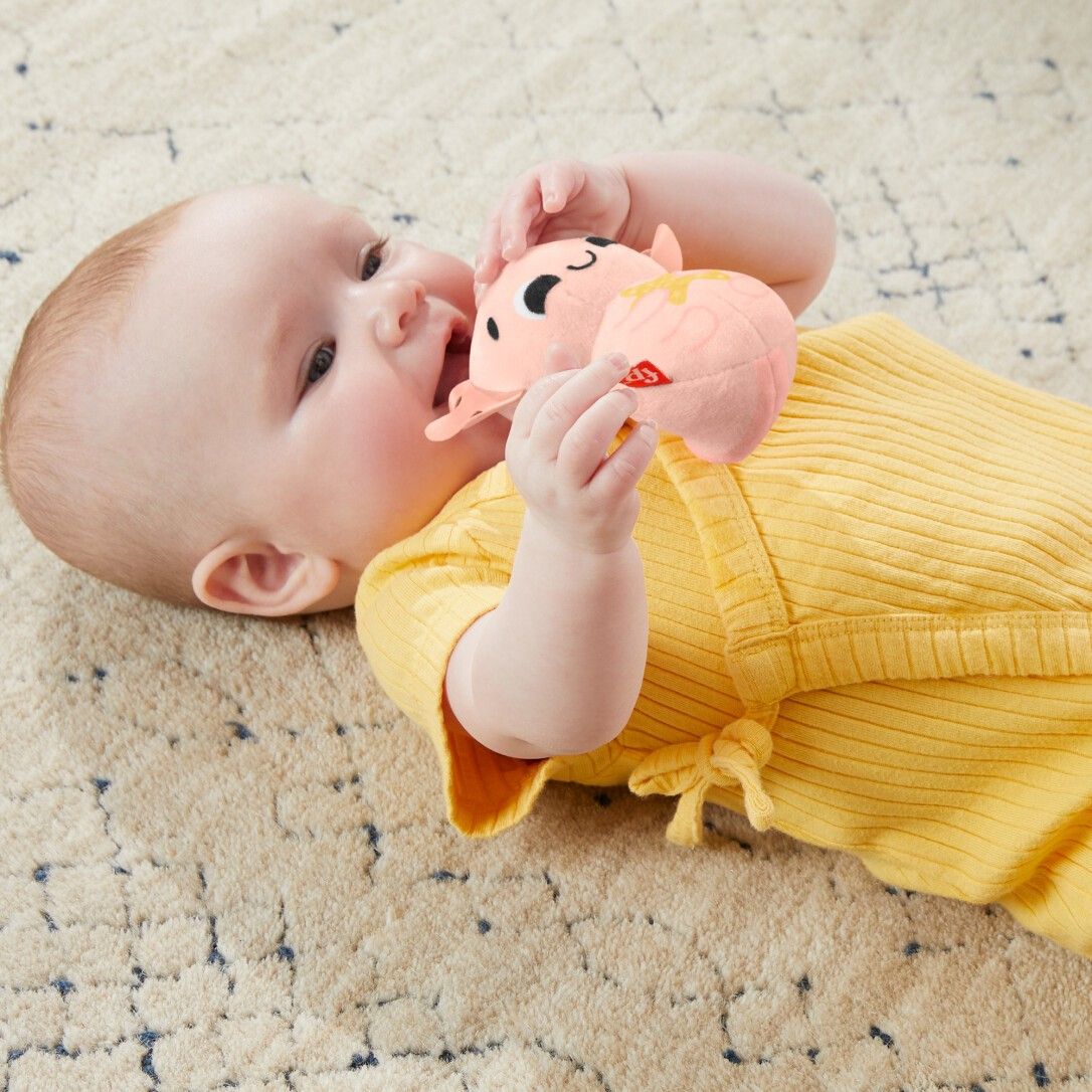 Baby liegt auf dem Bauch und hält ein rosa Spielzeug in Form eines Tieres. Das Spielzeug hat einen roten Aufnäher.