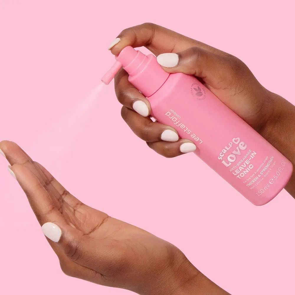 Hände halten eine rosa Sprühflasche. Produktname: Leave-In Tonic. Marke: Lee Stafford. Sprühnebel sichtbar. Vegan-Logo.