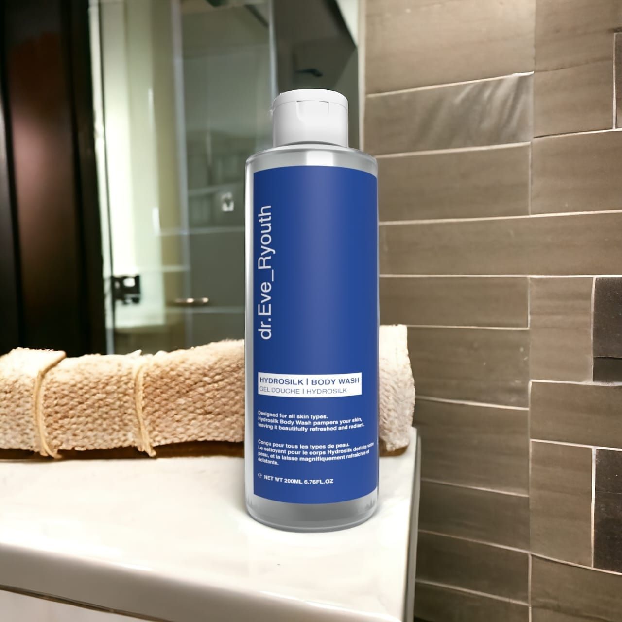 Klarflasche mit blauem Etikett. Aufschrift: dr.Eve_Ryouth, Hydrosilk Body Wash. Auf einer weißen Oberfläche, Handtuch im Hintergrund.