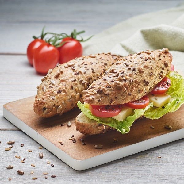 Zwei Körnerstangen, glutenfrei, auf einem Holzbrett. Aufgeschnitten mit Salat, Tomaten und Käse. Tomaten im Hintergrund.